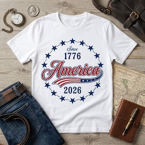 May include: A white short-sleeve t-shirt featuring a circular graphic with blue stars, the text "Since 1776", "America" in red script, and "2026" in blue, with a stylized American flag swoosh underneath the word America.