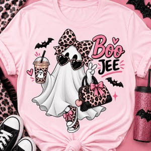 May include: A light pink t-shirt featuring a graphic of a white ghost wearing heart-shaped sunglasses, a leopard print bow, and leopard print sneakers. The ghost holds a leopard print handbag and a drink, with the text Boo JEE printed in pink and black.