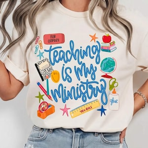 May include: A cream-coloured t-shirt featuring the phrase "Teaching is my Ministry" in blue script. The design incorporates school-related imagery, including an apple, books, a globe, and a pencil. This t-shirt is a casual, everyday wear item.