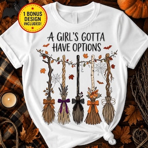 Puede incluir: Una camiseta blanca con el texto A GIRL'S GOTTA HAVE OPTIONS y una ilustración de cinco escobas distintas colgadas de una rama con hojas de otoño, telarañas y murciélagos. Una insignia amarilla indica 1 BONUS DESIGN INCLUDED.