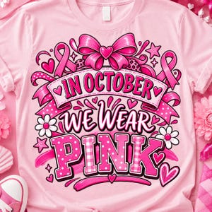 Puede incluir: Una camiseta rosa claro con un diseño gráfico que incluye el texto "IN OCTOBER WE WEAR PINK". El diseño presenta lazos rosas, corazones, estrellas y flores, con un gran lazo rosa en la parte superior. Las letras combinan patrones de lunares y cuadros.