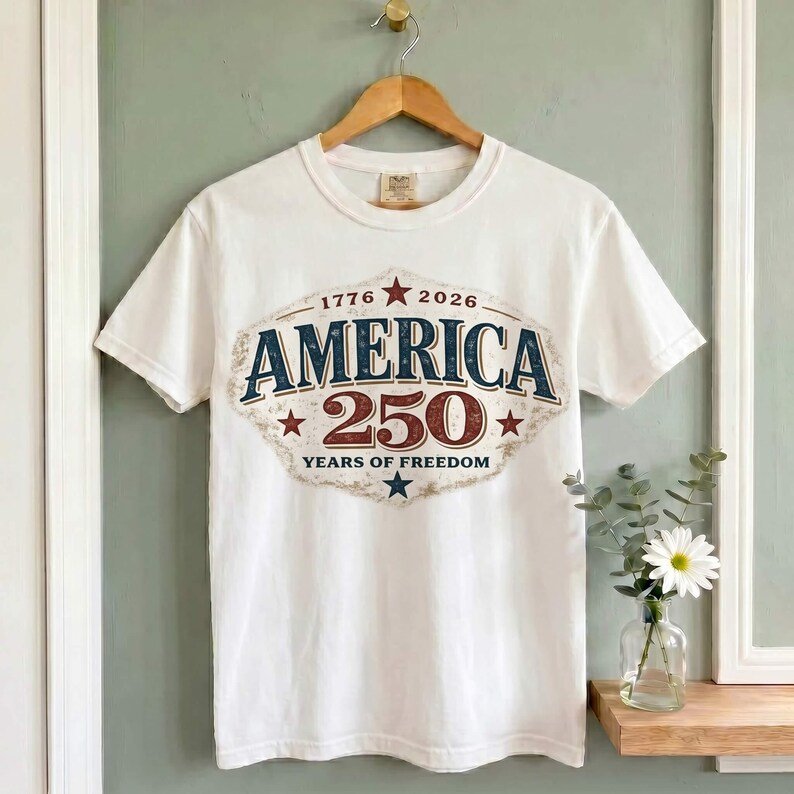 Pu&ograve; includere: T-shirt bianca con una grafica che commemora i 250 anni di libert&agrave; americana. Il design presenta la parola "AMERICA" in blu navy, il numero "250" in bordeaux e gli anni "1776" e "2026".