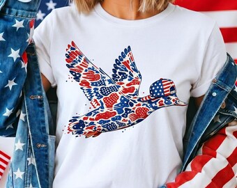 Può includere: T-shirt bianca con un design di anatra patriottica. L'anatra è in volo, con le ali spiegate, ed è colorata in motivi rossi, bianchi e blu. L'anatra indossa un berretto da baseball con il design della bandiera americana.