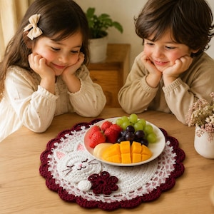 Puede incluir: Un tapete de ganchillo redondo en blanco y rojo oscuro con un diseño central de cara de gato, bigotes bordados y orejas rosas, acentuado por una aplicación de flor roja oscura. El tapete sirve como base para un plato con fruta fresca.