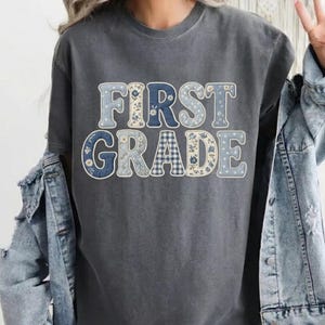 May include: A charcoal gray short-sleeve t-shirt featuring the text FIRST GRADE in large, block-style letters. Each letter is filled with various blue and white patterns, including floral prints, gingham checks, and polka dots, layered over a denim jacket.