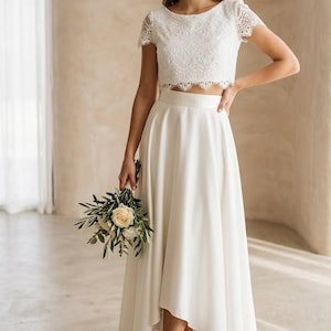 May include: A two-piece white outfit consisting of a short-sleeved lace crop top with a scalloped hem and a high-waisted, floor-length skirt featuring a high-low hemline. The skirt has a smooth, solid fabric finish and a clean, minimalist silhouette.