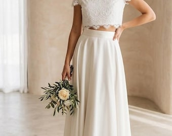 May include: A two-piece white outfit consisting of a short-sleeved lace crop top with a scalloped hem and a high-waisted, floor-length skirt featuring a high-low hemline. The skirt has a smooth, solid fabric finish and a clean, minimalist silhouette.