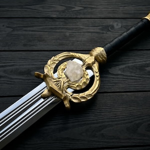 Puede incluir: Una espada con una hoja plateada pulida con múltiples ranuras, una guarda de metal dorado con un diseño ornamentado de hojas, un cristal transparente incrustado en el centro y un mango negro con un pomo dorado en el extremo.