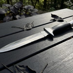May include: A fixed-blade knife featuring a double-edged, pointed steel blade with a serrated section near the hilt. The handle is black with a metallic pommel and a distinct crossguard separating the blade from the grip, creating a balanced design.