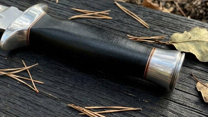 May include: A fixed-blade knife handle featuring a smooth black grip, polished silver-toned metal bolsters at both ends, and thin copper-colored accent rings separating the handle sections from the metallic components.