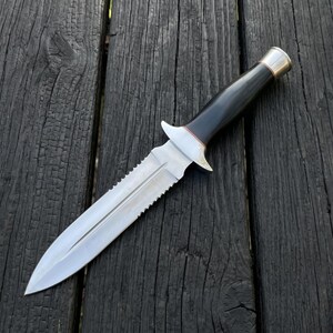 May include: A fixed-blade knife featuring a double-edged silver steel blade with a serrated spine, a silver metal crossguard, a smooth black handle, and a polished silver metal pommel cap at the end of the handle, creating a balanced tool design.