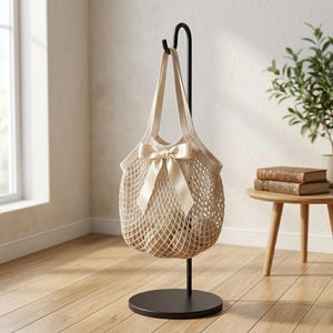 May include: A beige net shopping bag with a cream-colored bow is suspended from a black metal stand. The bag is made of a woven material and has a round base. The stand is on a wooden floor.