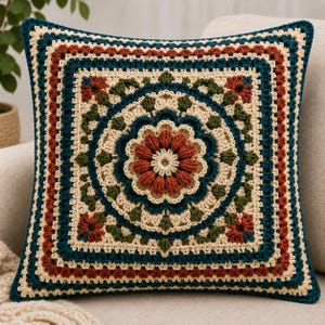 May include: A square crochet cushion cover featuring a central floral motif in rust orange, surrounded by concentric layers of cream, dark teal, and forest green yarn. The intricate design includes textured stitches and a repeating geometric border.