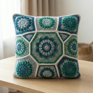 May include: A square crochet pillow cover featuring a geometric pattern of interconnected hexagonal and triangular motifs. The design uses a color palette of grey, teal, and dark blue yarn, with cream-colored borders outlining each shape.