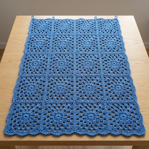 Puede incluir: Una manta azul tejida a ganchillo con un diseño floral, exhibida sobre una mesa de madera. La manta está hecha de cuadrados unidos, con un borde festoneado y lazos para colgar. El color es un azul vibrante y medio.