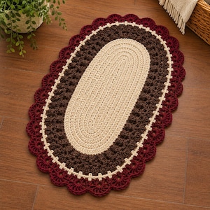 May include: An oval-shaped crochet rug featuring a central cream-colored section, a surrounding dark brown textured border, and an outer scalloped edge in deep burgundy. The rug is constructed with visible yarn stitches and a consistent pattern throughout.