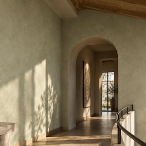 May include: A beige stone console table with two cylindrical bases, topped with a large ceramic vase containing leafy branches, a small bowl, and a stack of books, positioned against a textured sage green wall in a hallway.