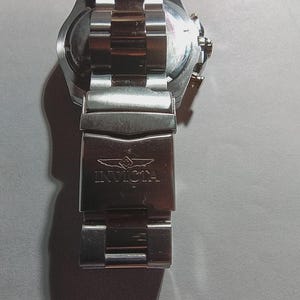 May include: A stainless steel Invicta watch with a silver and gold band. The watch face is visible, and the clasp is engraved with the Invicta logo. The watch band is approximately 8 inches long.