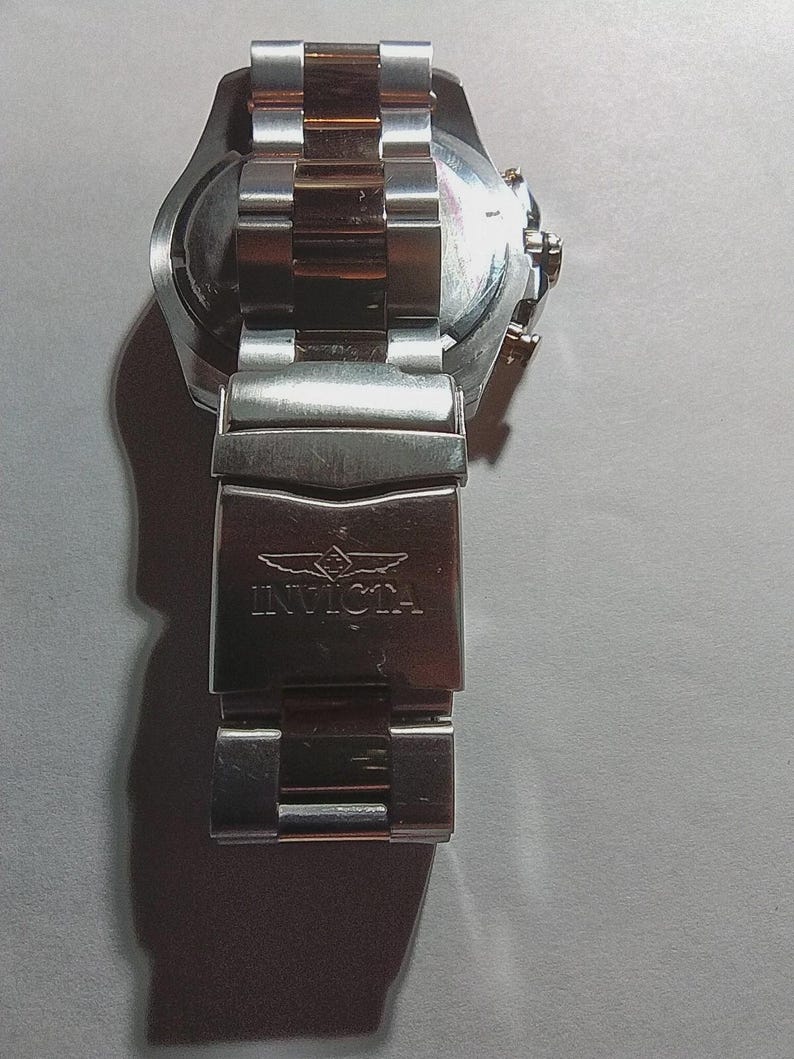 May include: A stainless steel Invicta watch with a silver and gold band. The watch face is visible, and the clasp is engraved with the Invicta logo. The watch band is approximately 8 inches long.