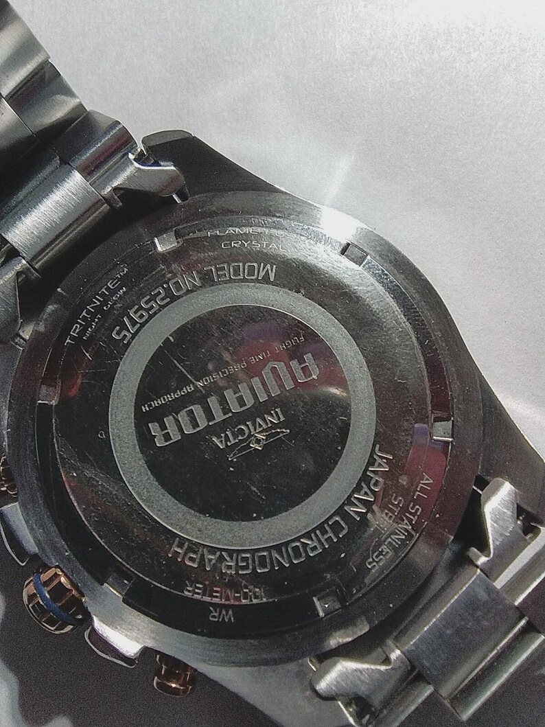 May include: Close-up of the back of a stainless steel Invicta Aviator chronograph watch. The watch features the text "AVIATOR" and "JAPAN CHRONOGRAPH". The watch is water resistant to 100 meters.