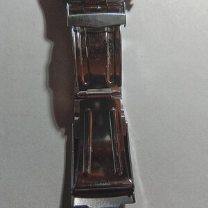 May include: Close-up of a silver stainless steel watch clasp. The clasp is open, revealing the inner mechanism. The watch band is also visible, with its shiny, metallic links. The watch appears to be new.