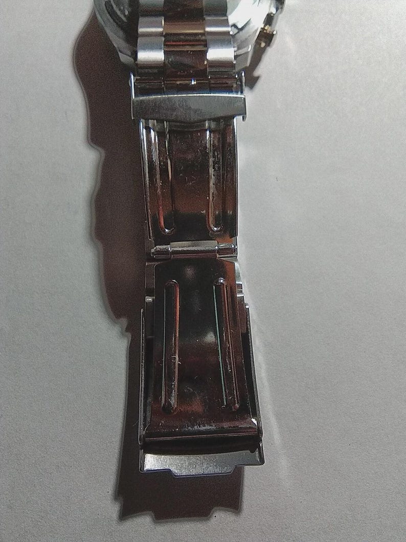 May include: Close-up of a silver stainless steel watch clasp. The clasp is open, revealing the inner mechanism. The watch band is also visible, with its shiny, metallic links. The watch appears to be new.