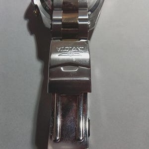 May include: Close-up of a silver and gold-toned Invicta watch clasp. The clasp is made of stainless steel and features the Invicta logo. The watch band is partially visible, showing a two-tone design.