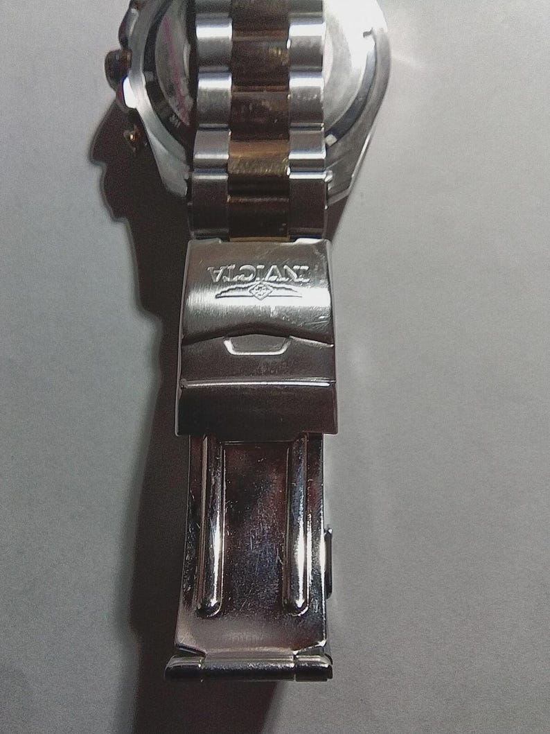 May include: Close-up of a silver and gold-toned Invicta watch clasp. The clasp is made of stainless steel and features the Invicta logo. The watch band is partially visible, showing a two-tone design.