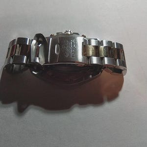 May include: A silver and gold Invicta watch with a stainless steel band. The watch clasp is open, revealing the Invicta logo. The watch band has silver and gold links.