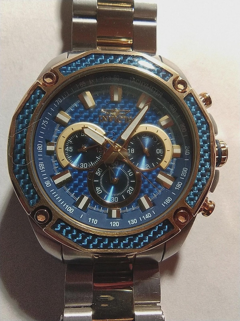 May include: A stainless steel Invicta watch with a blue and gold face. The watch features a blue carbon fiber bezel and three sub-dials. The watch has gold accents and a stainless steel band.