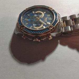 May include: A stainless steel wristwatch with a blue face and gold accents. The watch features a blue bezel and a silver and gold band. The watch has three sub-dials and gold hands. The watch is a chronograph style.