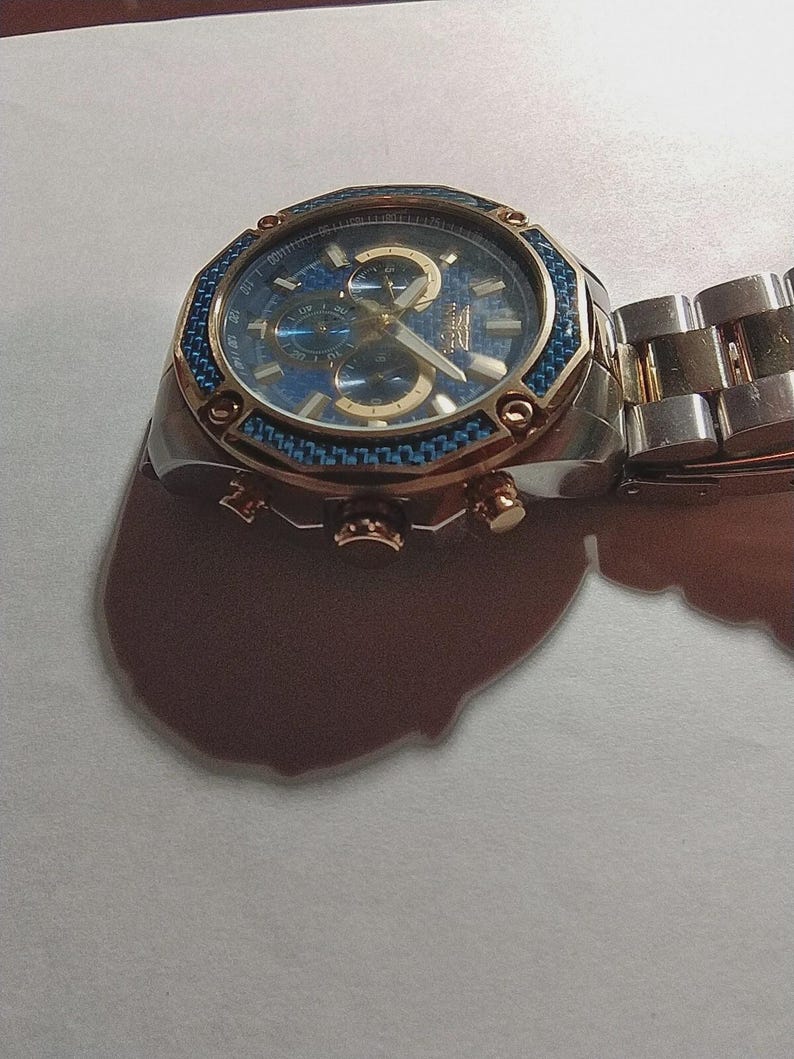 May include: A stainless steel wristwatch with a blue face and gold accents. The watch features a blue bezel and a silver and gold band. The watch has three sub-dials and gold hands. The watch is a chronograph style.