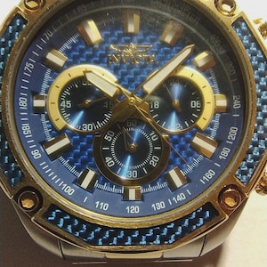 May include: Close-up of an Invicta watch with a blue and gold design. The watch features a blue face with gold accents, three sub-dials, and a gold bezel. The watch band is silver. The watch has a sporty, luxury aesthetic.