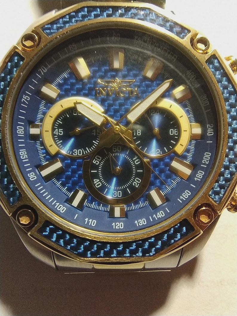 May include: Close-up of an Invicta watch with a blue and gold design. The watch features a blue face with gold accents, three sub-dials, and a gold bezel. The watch band is silver. The watch has a sporty, luxury aesthetic.