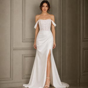 Puede incluir: Un vestido blanco largo hasta el suelo con escote de hombros descubiertos, un corpiño de encaje texturizado y una falda de satén suave. La falda incluye una abertura lateral alta que deja ver la pierna, creando una silueta drapeada hasta el suelo.