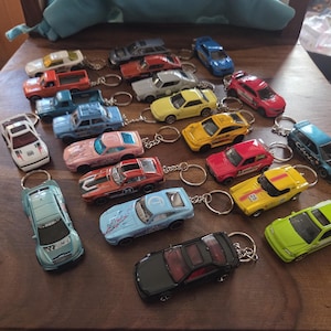 Könnte beinhalten: Eine Sammlung verschiedener Spielzeugauto-Schlüsselanhänger aus Druckguss in verschiedenen Farben wie Rot, Blau, Gelb, Grün, Rosa, Weiß und Schwarz. Jedes kleine Fahrzeug hat einen Metallring am Dach befestigt.