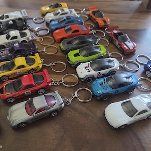 Könnte beinhalten: Eine Sammlung verschiedener Spielzeugauto-Schlüsselanhänger in vielen Farben wie Gelb, Rot, Weiß, Blau, Schwarz, Silber, Orange, Lila und Grün. Jedes Miniaturfahrzeug verfügt über einen Metallring am Dach oder Heck und zeigt diverse Sportwagen-Designs und Rennlackierungen.