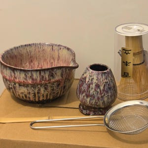 Könnte beinhalten: Eine Keramik-Matchaschale und ein Besenhalter mit violetter und weißer Tropfglasur, ein Bambus-Teelöffel, ein Metallsieb und ein Bambusbesen in einem transparenten zylindrischen Behälter mit den japanischen Schriftzeichen 本立 auf einem Goldetikett.