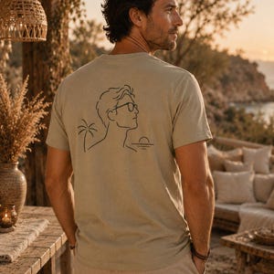 Op de afbeelding: Een lichtbeige t-shirt met korte mouwen en een zwarte lijntekening op de achterkant. Het ontwerp toont het profiel van een persoon met een bril, naast een palmboom en een ondergaande zon boven een horizonlijn.
