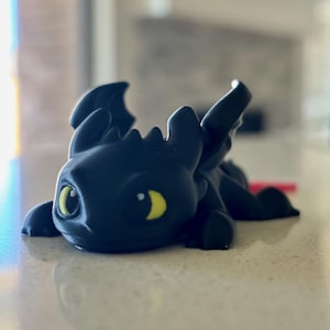 May include: A black dragon figurine with yellow eyes and small wings. The dragon is lying down on a light-colored surface. The figurine has a smooth texture and is likely 3D-printed.