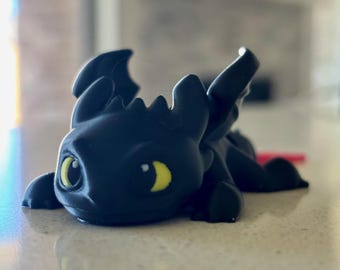 May include: A black dragon figurine with yellow eyes and small wings. The dragon is lying down on a light-colored surface. The figurine has a smooth texture and is likely 3D-printed.