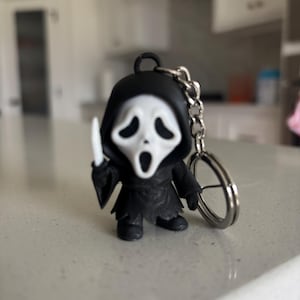 May include: A small, stylized keychain figure of a character wearing a black hooded robe and a white mask with a wide, open-mouthed expression. The figure holds a small white knife in its right hand and is attached to a silver metal keyring.