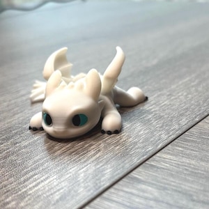 May include: A white dragon figurine with large, teal eyes and black claws. The dragon is lying down with its wings spread. The figurine is made of a smooth material and is a fantasy-themed collectible.