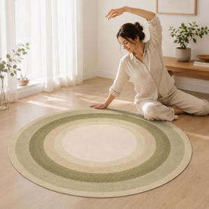 May include: A circular area rug featuring a concentric ring pattern in shades of sage green, olive green, and cream. The rug has a flat-woven texture and is positioned on a light-coloured wooden floor, showcasing a gradient of earthy tones.