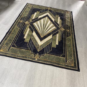 May include: A rectangular area rug featuring a dark navy blue, olive green, and cream geometric pattern. The design includes a central diamond shape with radiating fan motifs, surrounded by layered angular lines and borders in gold and black.