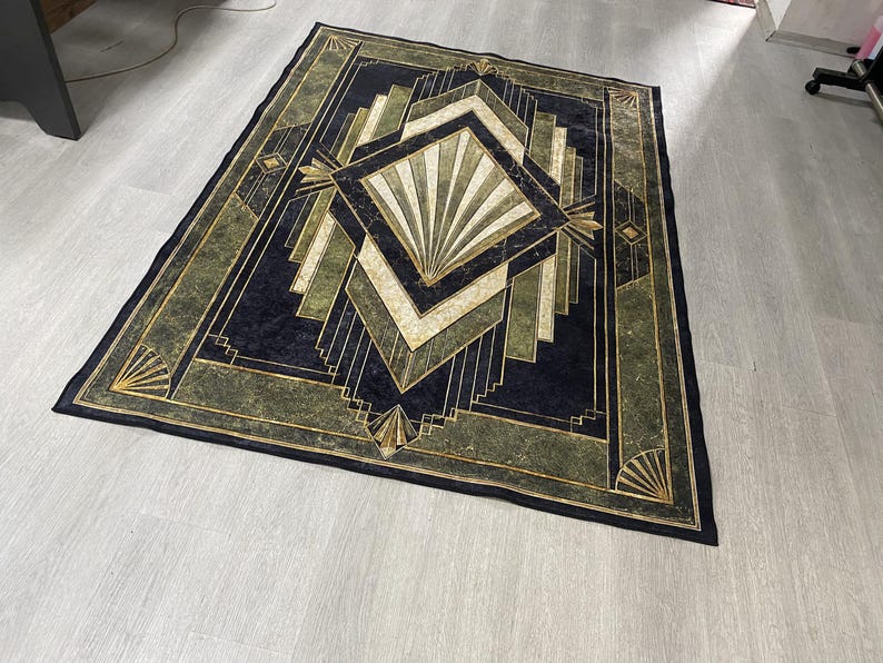 May include: A rectangular area rug featuring a dark navy blue, olive green, and cream geometric pattern. The design includes a central diamond shape with radiating fan motifs, surrounded by layered angular lines and borders in gold and black.