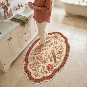May include: An oval-shaped bathroom rug featuring a cream-coloured centre with a detailed floral pattern of pink peonies and two brown birds perched on branches. The rug has a wide, dark red scalloped border with intricate decorative motifs.