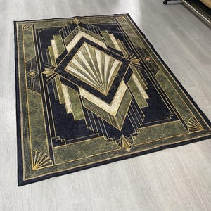 May include: A rectangular area rug featuring a dark navy blue and olive green geometric pattern. The design includes a central diamond shape with radiating lines, surrounded by layered angular borders and fan-like motifs in gold and cream tones.