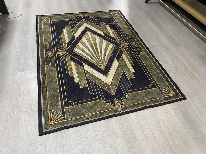 May include: A rectangular area rug featuring a dark navy blue and olive green geometric pattern. The design includes a central diamond shape with radiating lines, surrounded by layered angular borders and fan-like motifs in gold and cream tones.