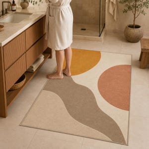May include: A rectangular bathroom rug featuring a graphic pattern of large, abstract shapes in mustard yellow, terracotta orange, and taupe on a cream background. The rug has a finished edge and is positioned on a light-colored tiled floor.